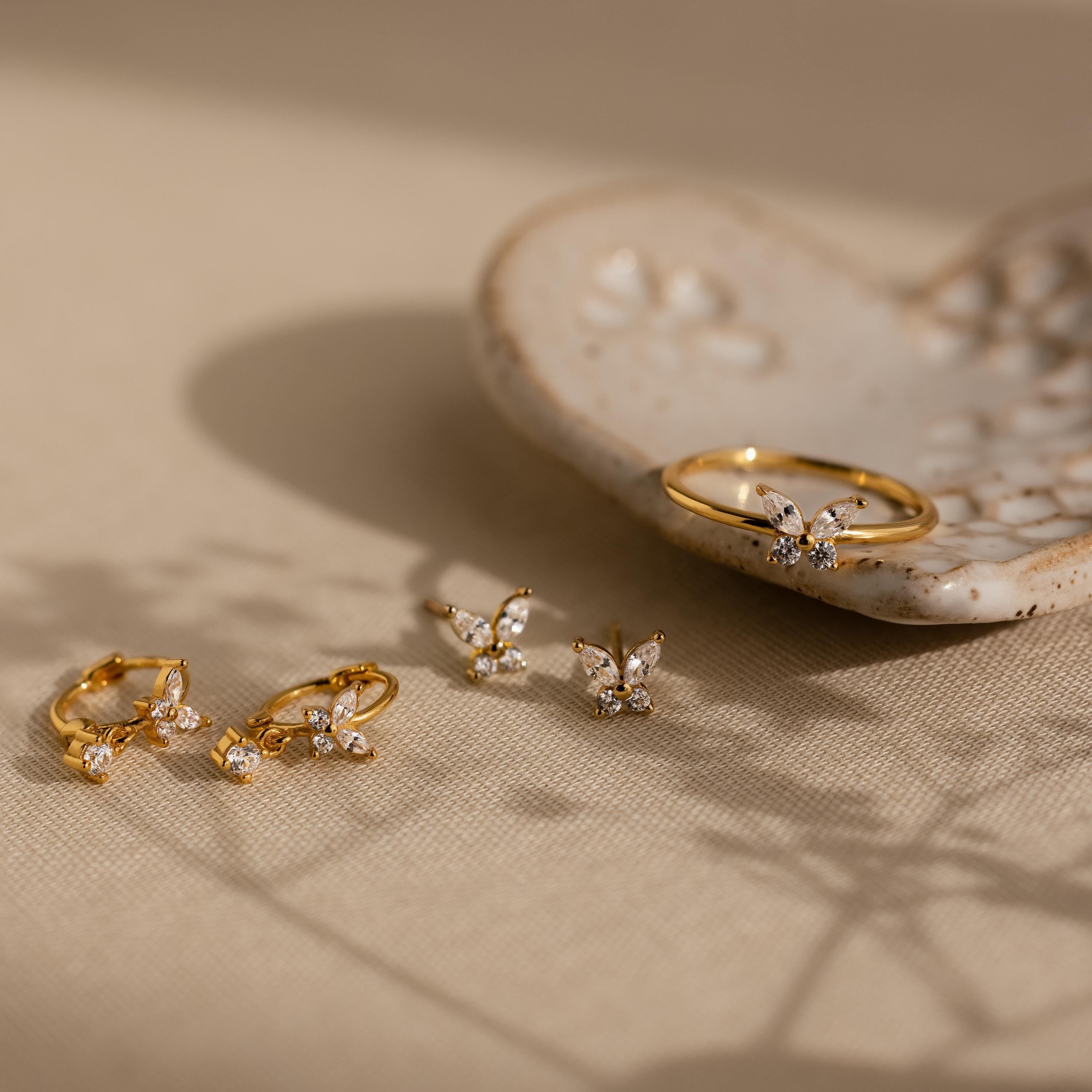 Butterfly Drop Huggies displayed on a beige surface with a ceramic dish in the background, featuring elegant butterfly-inspired earrings accented with sparkling diamonds.
