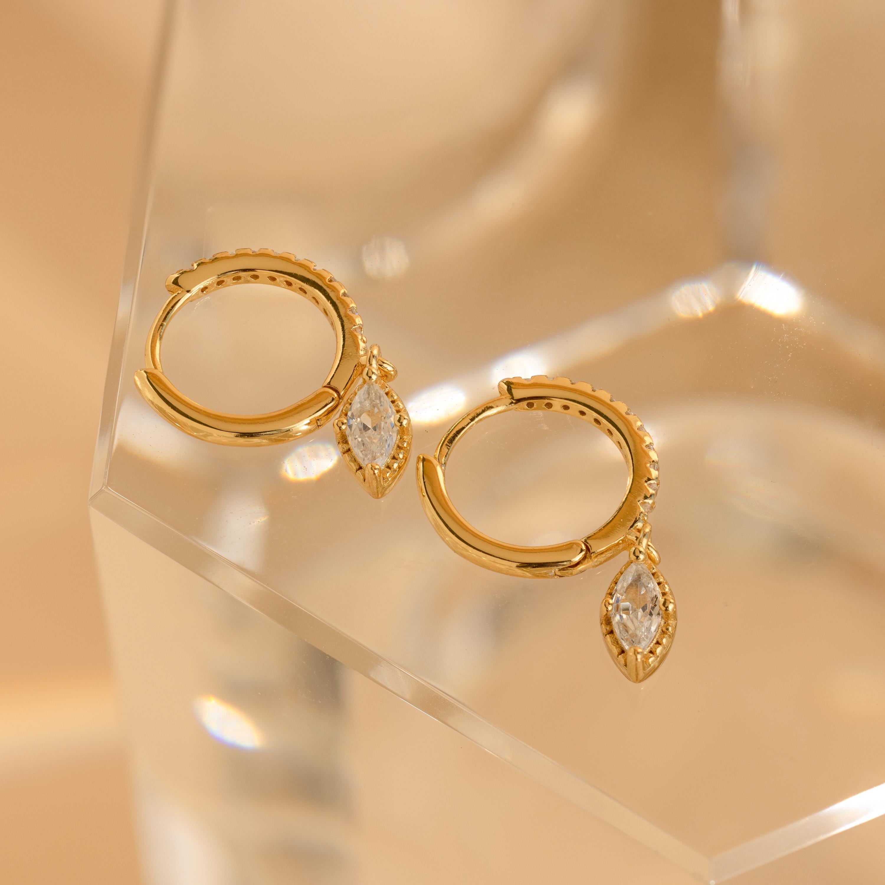 Marquise Pave Drop Huggies: Elegant gold hoop earrings feature marquise-cut crystal pendants, capturing the grace of marquise drop huggies and shimmering beautifully on a clear, faceted surface.