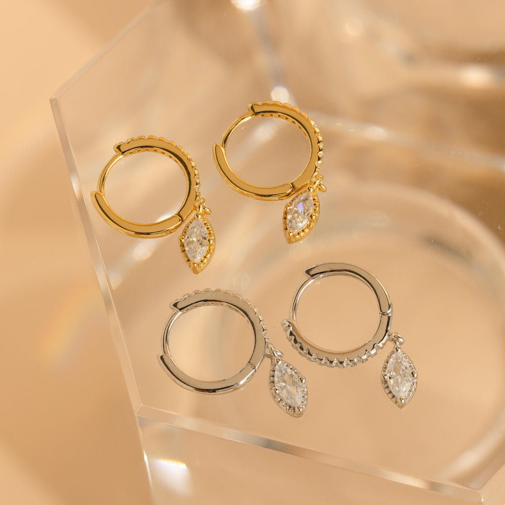 The Marquise Pave Drop Huggies set includes two pairs of elegant earrings, one in gold and one in silver, each adorned with a dangling marquise crystal charm for a sophisticated touch to any outfit.