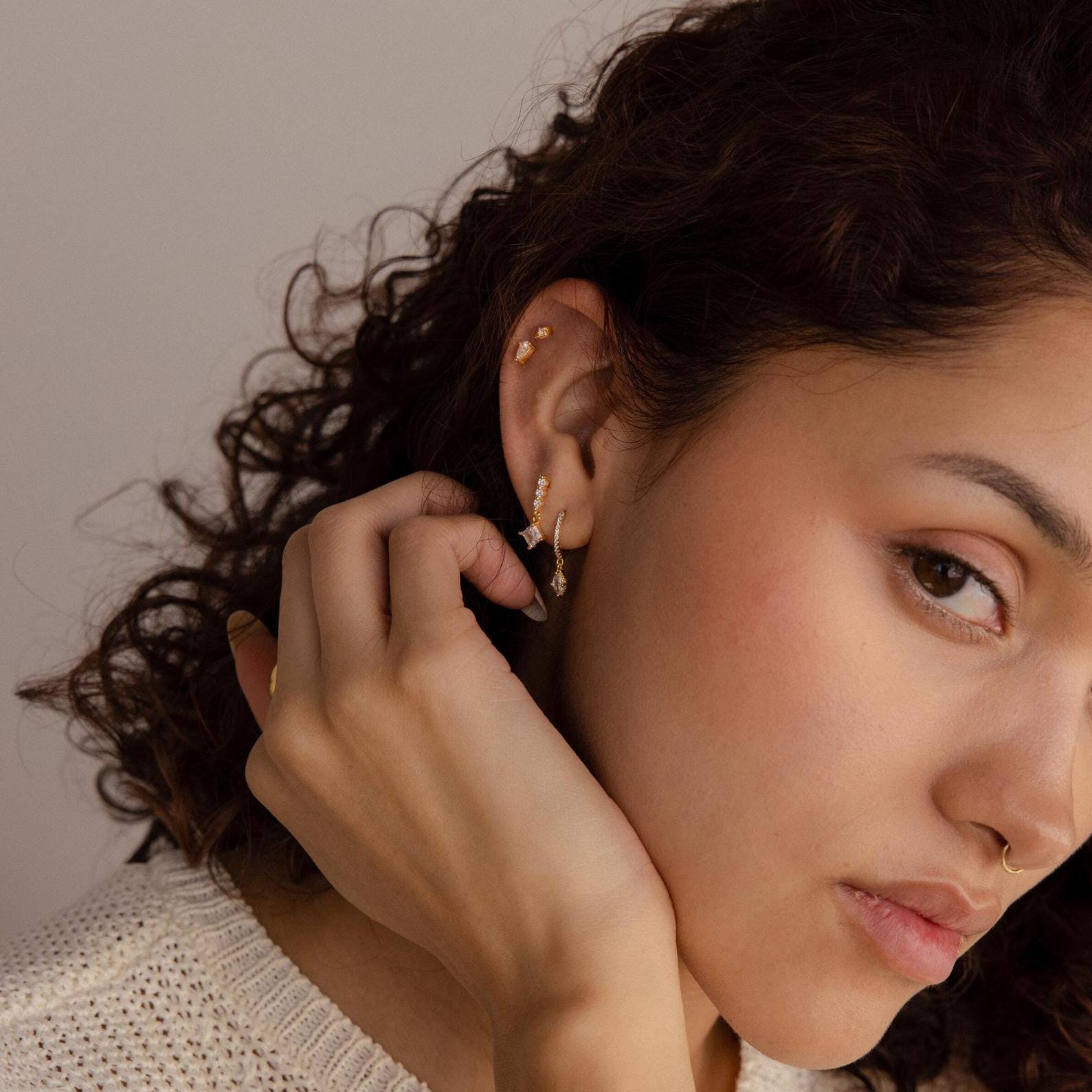 A woman with curly hair wearing Marquise Pave Drop Huggies and a cream sweater gently touches her ear.