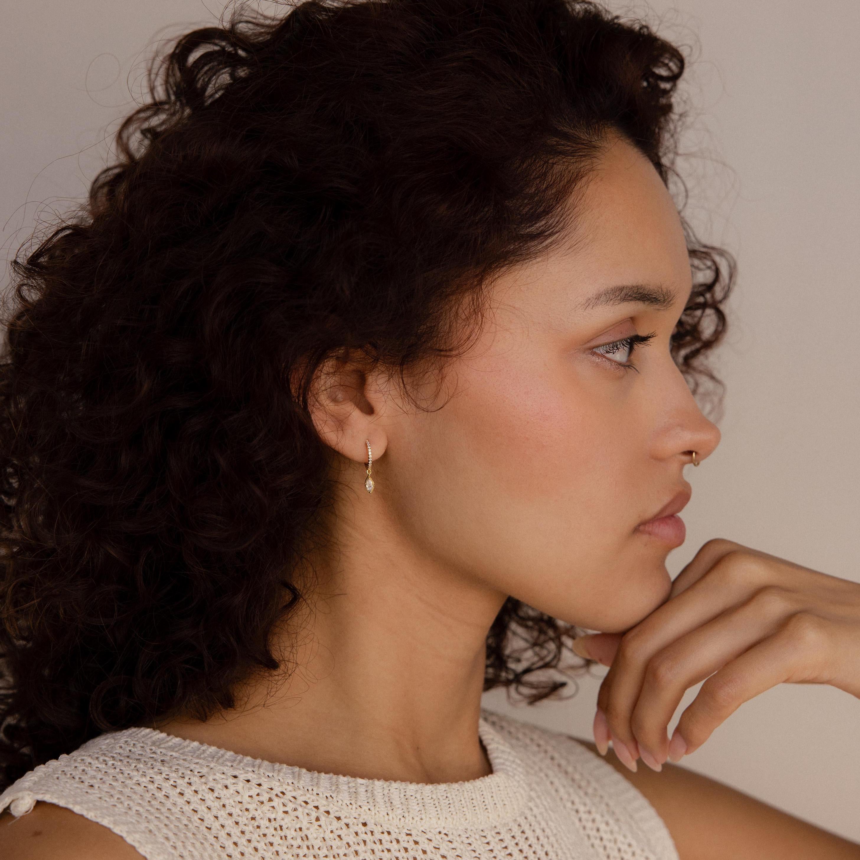 A woman with curly hair in profile, wearing a white knit top, elegantly showcases the Marquise Pave Drop Huggies as she rests her chin on her hand.