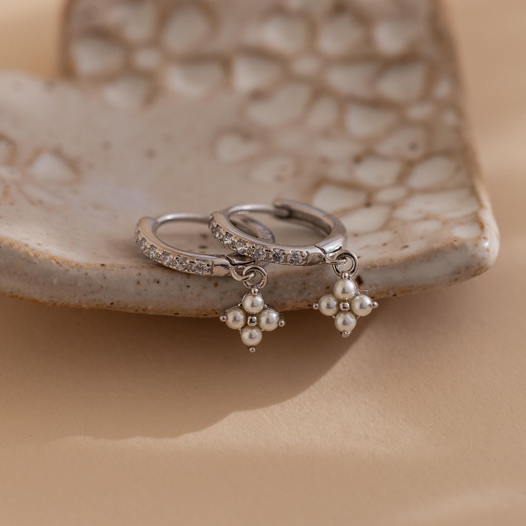 The Pearl Ivy Flower Huggies in Sterling Silver feature two silver hoop earrings with pearl drops and crystal accents, displayed on a textured ceramic dish to add a delicate floral touch to your jewelry collection.
