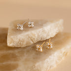 Two pairs of stud earrings, including Pearl Ivy Flower Studs in silver and gold, displayed on beige stone slabs.