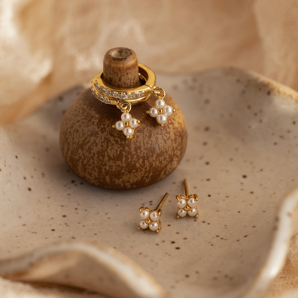 The Pearl Ivy Flower Studs in 18K Gold are displayed on a rustic brown holder with a beige ceramic dish background—an elegant choice for wedding jewelry.