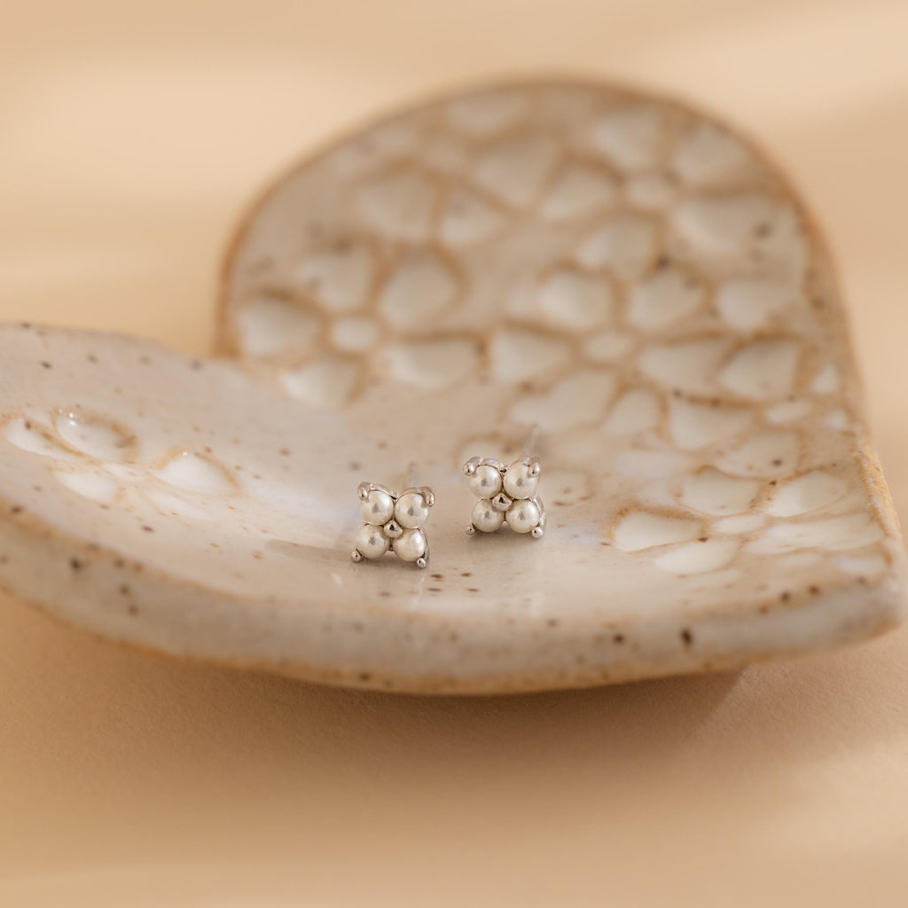 The Pearl Ivy Flower Studs are small silver floral earrings displayed on a heart-shaped, textured ceramic dish against a soft beige background.