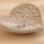 The Pearl Ivy Flower Studs are small silver floral earrings displayed on a heart-shaped, textured ceramic dish against a soft beige background.