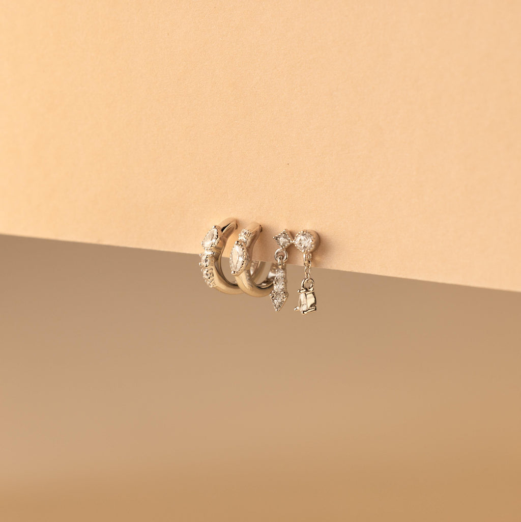 The Dainty Pave Earrings Set, featuring three gold hoop earrings and a dangling diamond-accented earring, is beautifully displayed on a beige background.