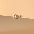 The Dainty Pave Earrings Set, featuring three gold hoop earrings and a dangling diamond-accented earring, is beautifully displayed on a beige background.