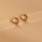 Dangling Pearl Diamond Huggies in 18K Gold rest on a beige surface, adorned with delicate pearls and sparkling diamonds, illuminated by faint rainbow light—perfect for lovers of elegant dangling pearl earrings.