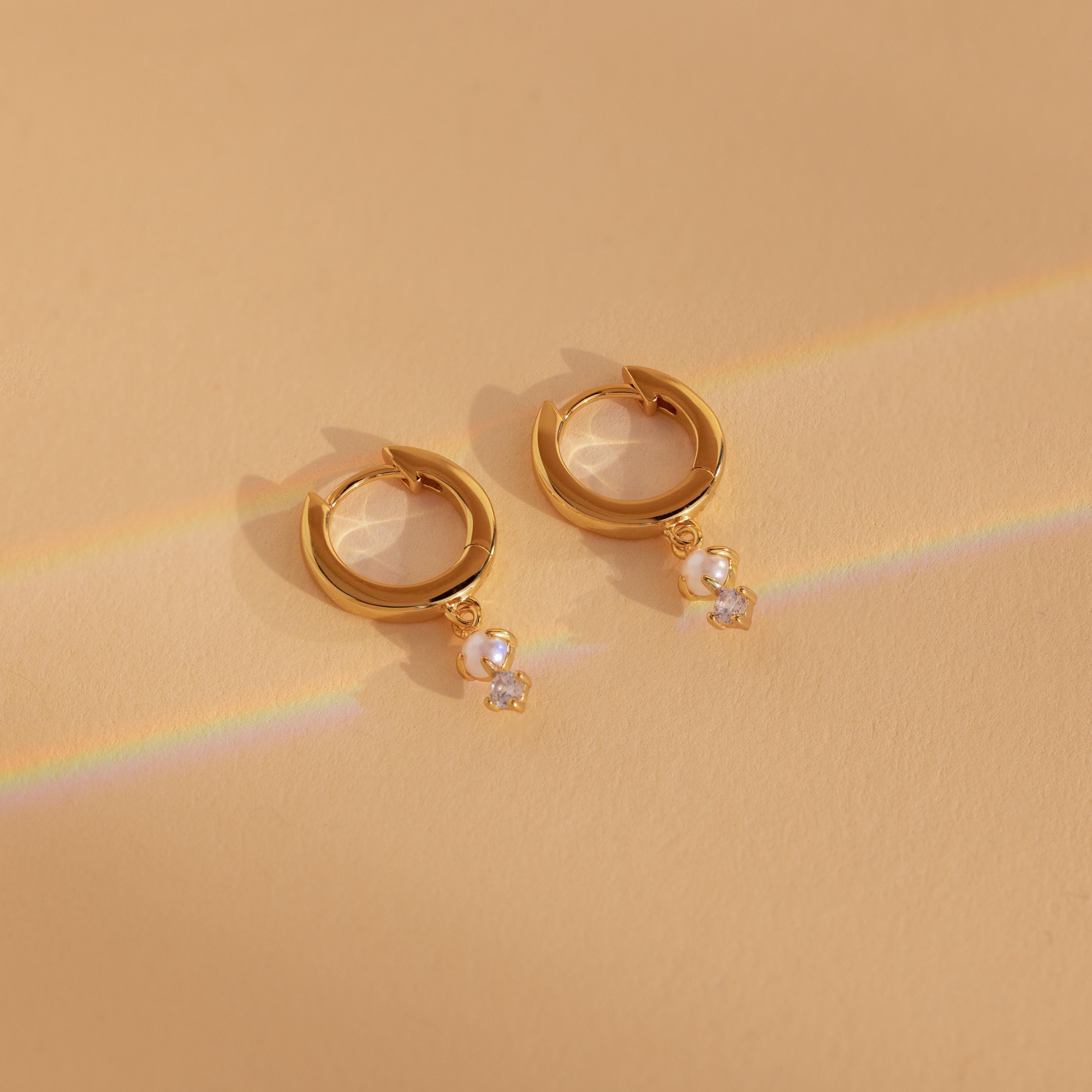 Dangling Pearl Diamond Huggies in 18K Gold rest on a beige surface, adorned with delicate pearls and sparkling diamonds, illuminated by faint rainbow light—perfect for lovers of elegant dangling pearl earrings.