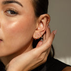 A woman with glowing skin touches her ear, wearing the Dainty Pave Earrings Set—multiple small gold hoops and studs that add subtle sparkle.