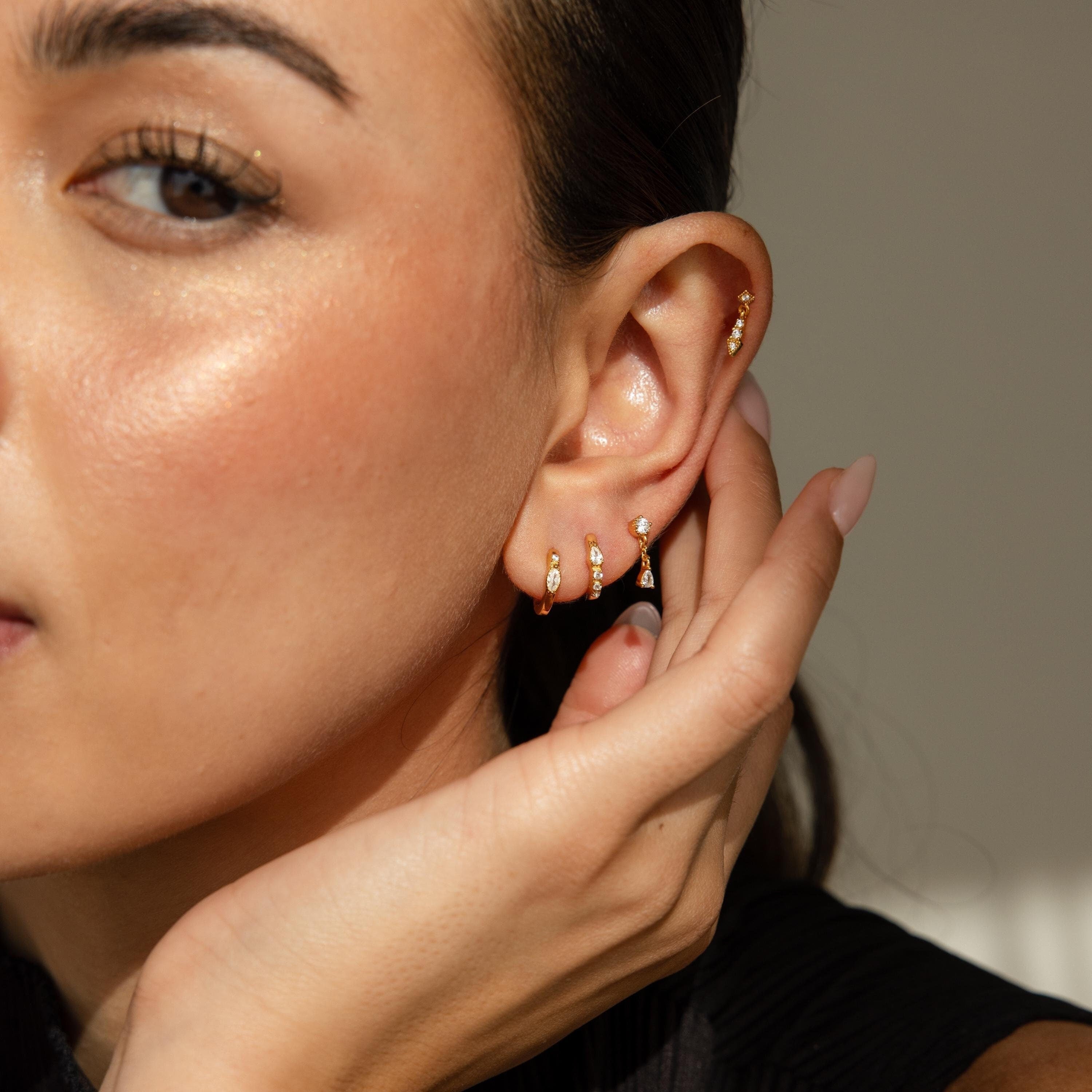 A woman with glowing skin touches her ear, wearing the Dainty Pave Earrings Set—multiple small gold hoops and studs that add subtle sparkle.