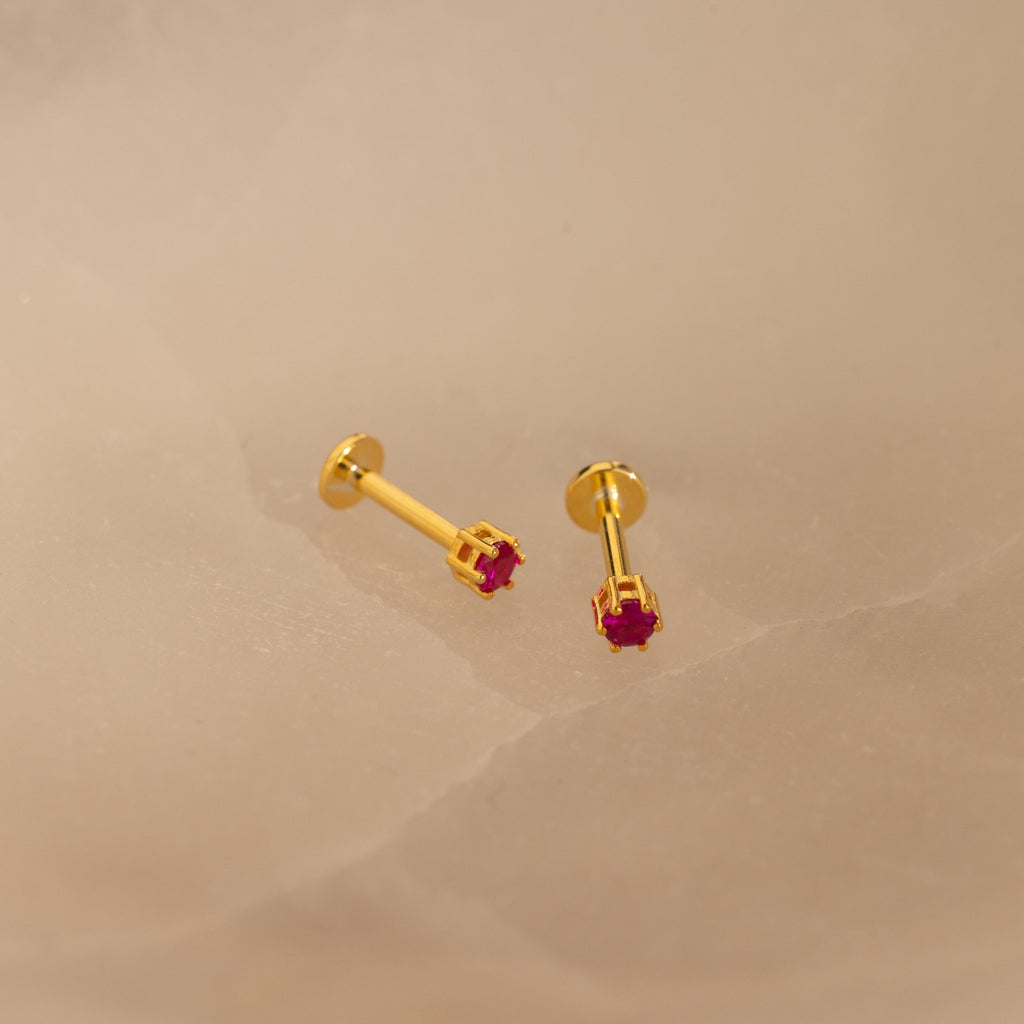 The Ruby Flatback Studs, featuring small red gemstones set in gold, rest on a light beige surface and add a subtle pop of color to your collection.