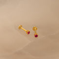 The Ruby Flatback Studs, featuring small red gemstones set in gold, rest on a light beige surface and add a subtle pop of color to your collection.