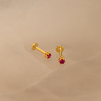 The Ruby Flatback Studs, featuring small red gemstones set in gold, rest on a light beige surface and add a subtle pop of color to your collection.