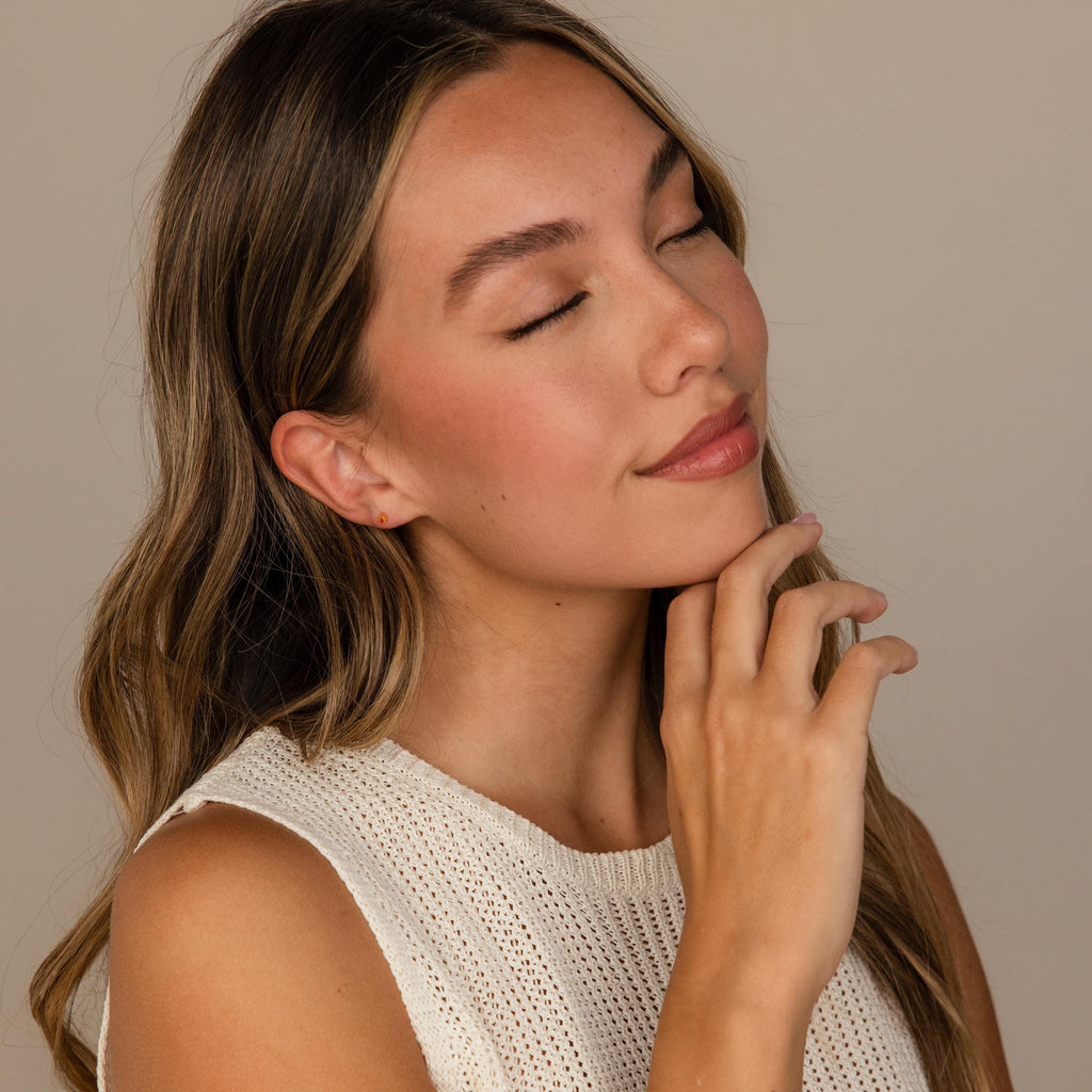 Woman with long brown hair and closed eyes gently touching her chin, wearing a sleeveless knit top and sparkling Ruby Flatback Studs.