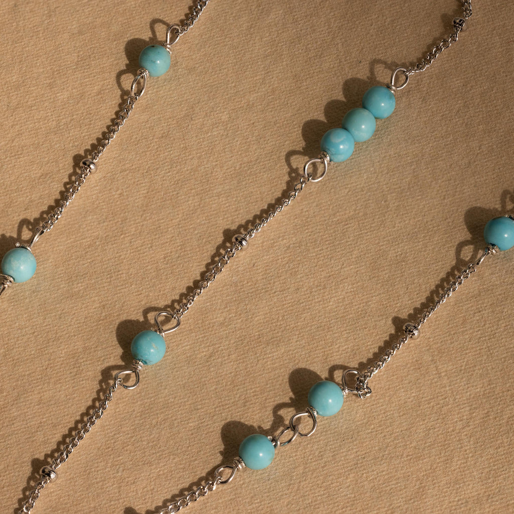 The Turquoise Satellite Necklace features silver chains with turquoise beads arranged diagonally on a textured beige surface, perfect for layering or wearing as a standout piece.