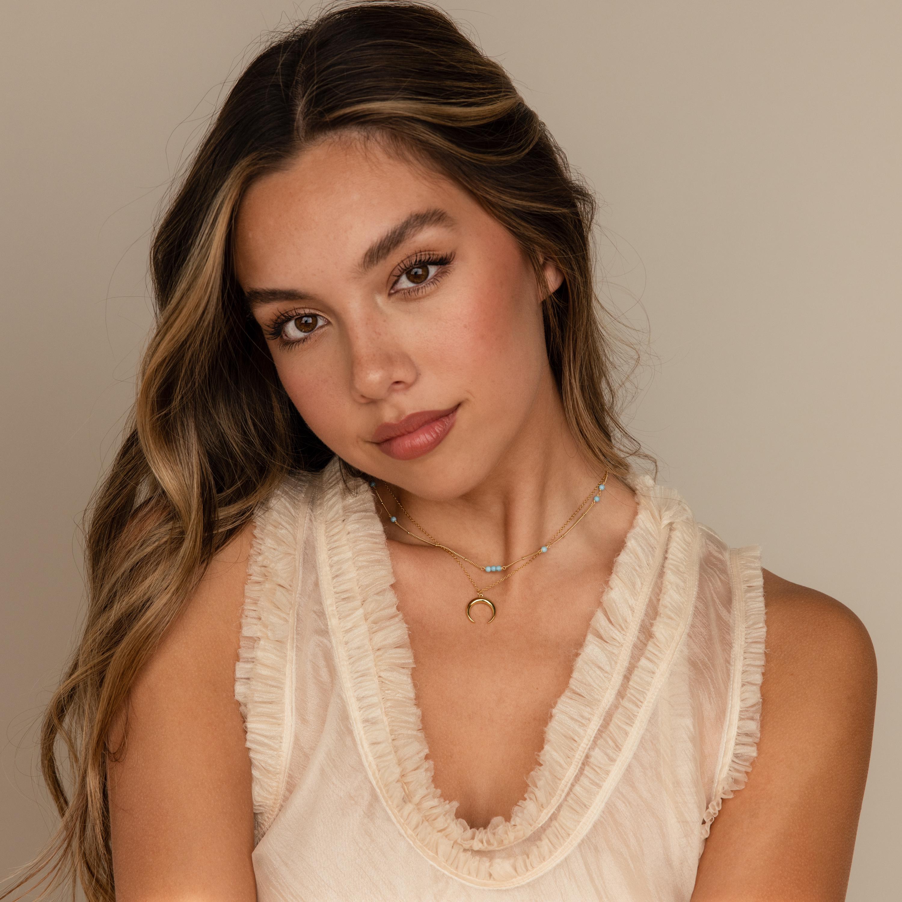 Young woman with long wavy hair wears a sleeveless cream ruffled top and the Turquoise Satellite Necklace, featuring turquoise beads, posing calmly.