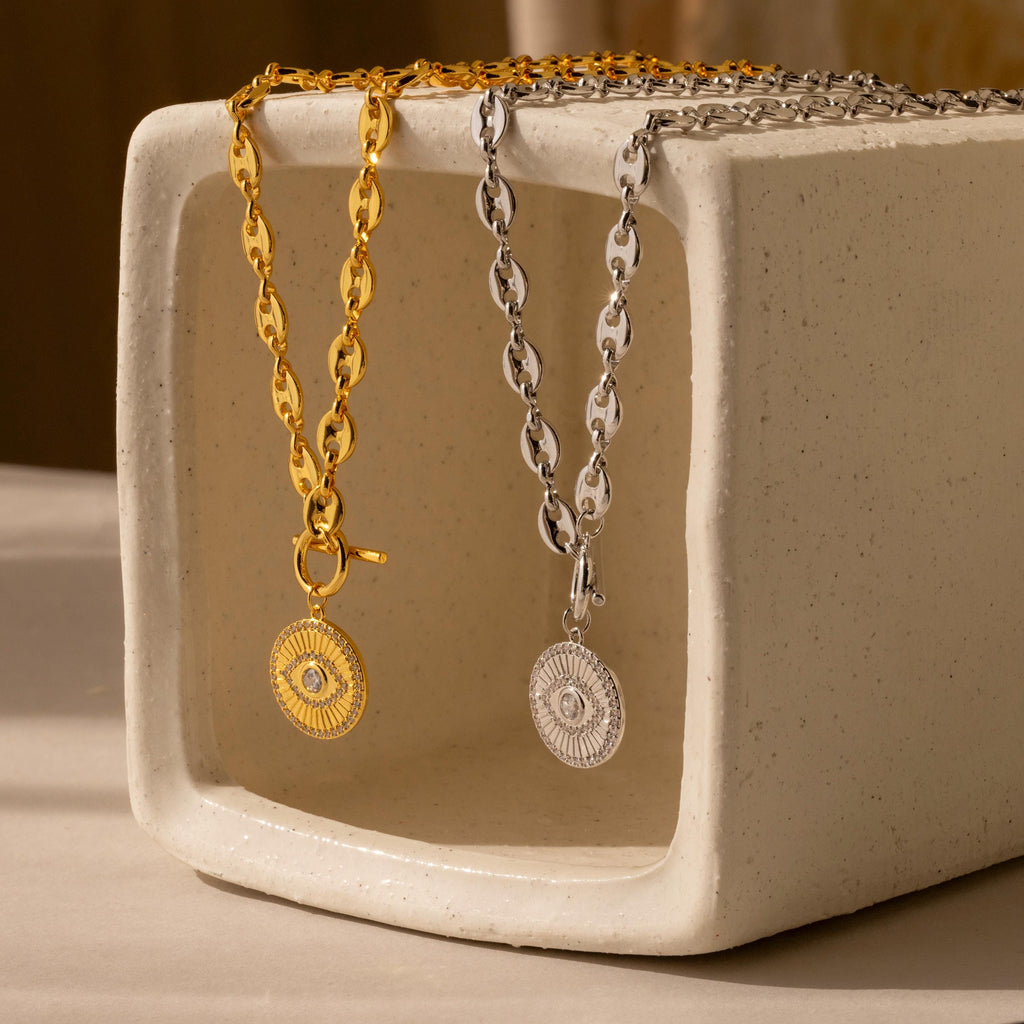 The Evil Eye Toggle Necklace features a gold and silver mariner chain with a striking diamond Evil Eye charm, displayed on a rectangular ceramic stand.