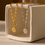The Evil Eye Toggle Necklace features a gold and silver mariner chain with a striking diamond Evil Eye charm, displayed on a rectangular ceramic stand.