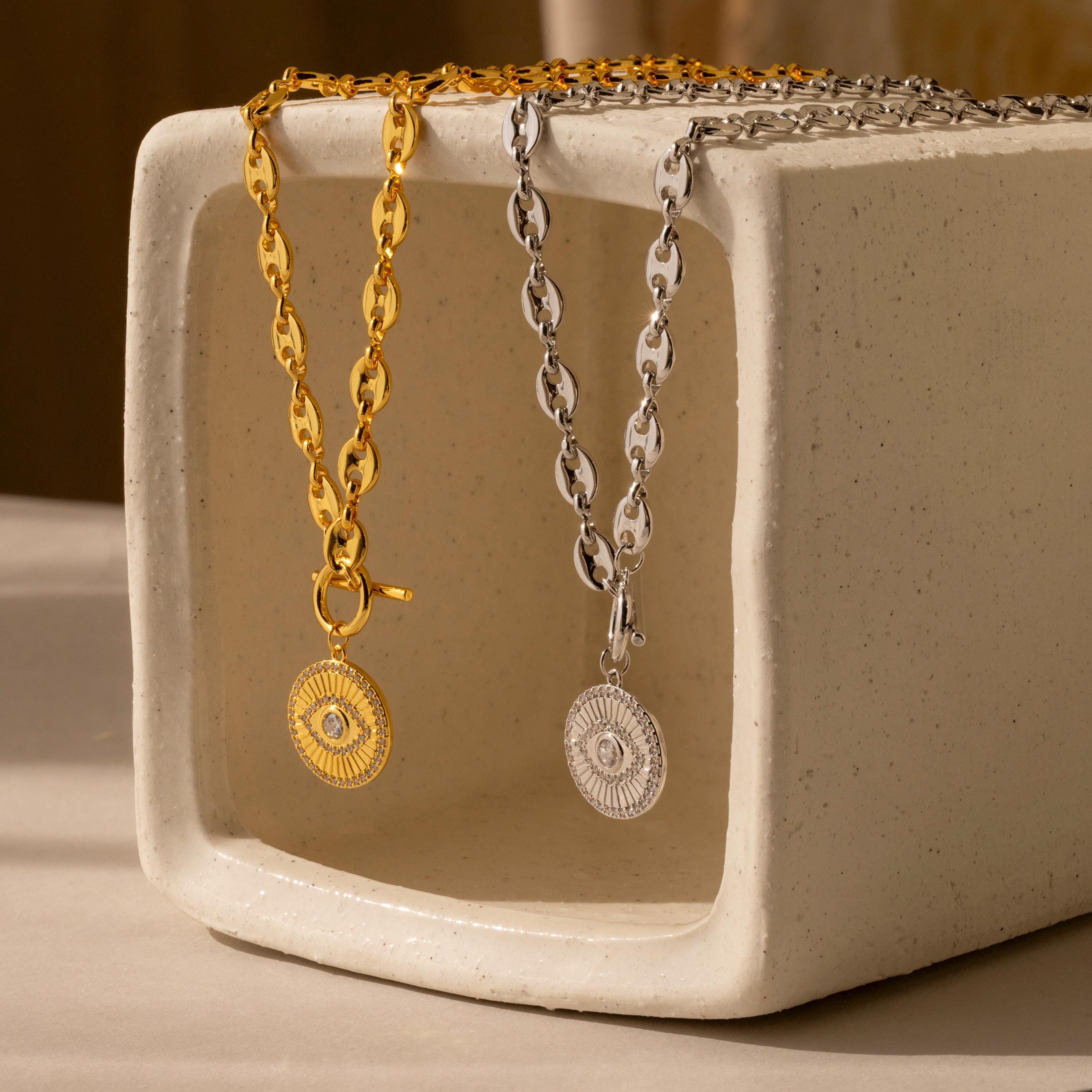 The Evil Eye Toggle Necklace features a gold and silver mariner chain with a striking diamond Evil Eye charm, displayed on a rectangular ceramic stand.