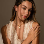 Young woman in a cream ruffled top wearing gold jewelry, including an Evil Eye Toggle Necklace, smiling softly in warm, natural light.