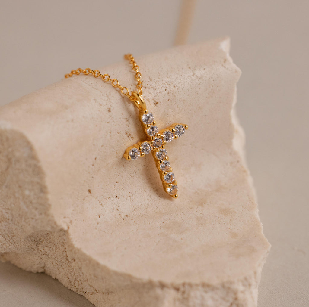 The Pave Cross Necklace features a gold chain and a diamond-studded cross pendant, elegantly displayed on a beige textured stone surface—a stunning piece of jewelry.
