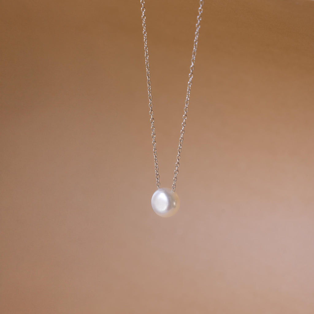 The Classic Pearl Pendant Necklace is a delicate gold chain with a single round pearl, showcased against a soft beige background.