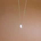 The Classic Pearl Pendant Necklace is a delicate gold chain with a single round pearl, showcased against a soft beige background.