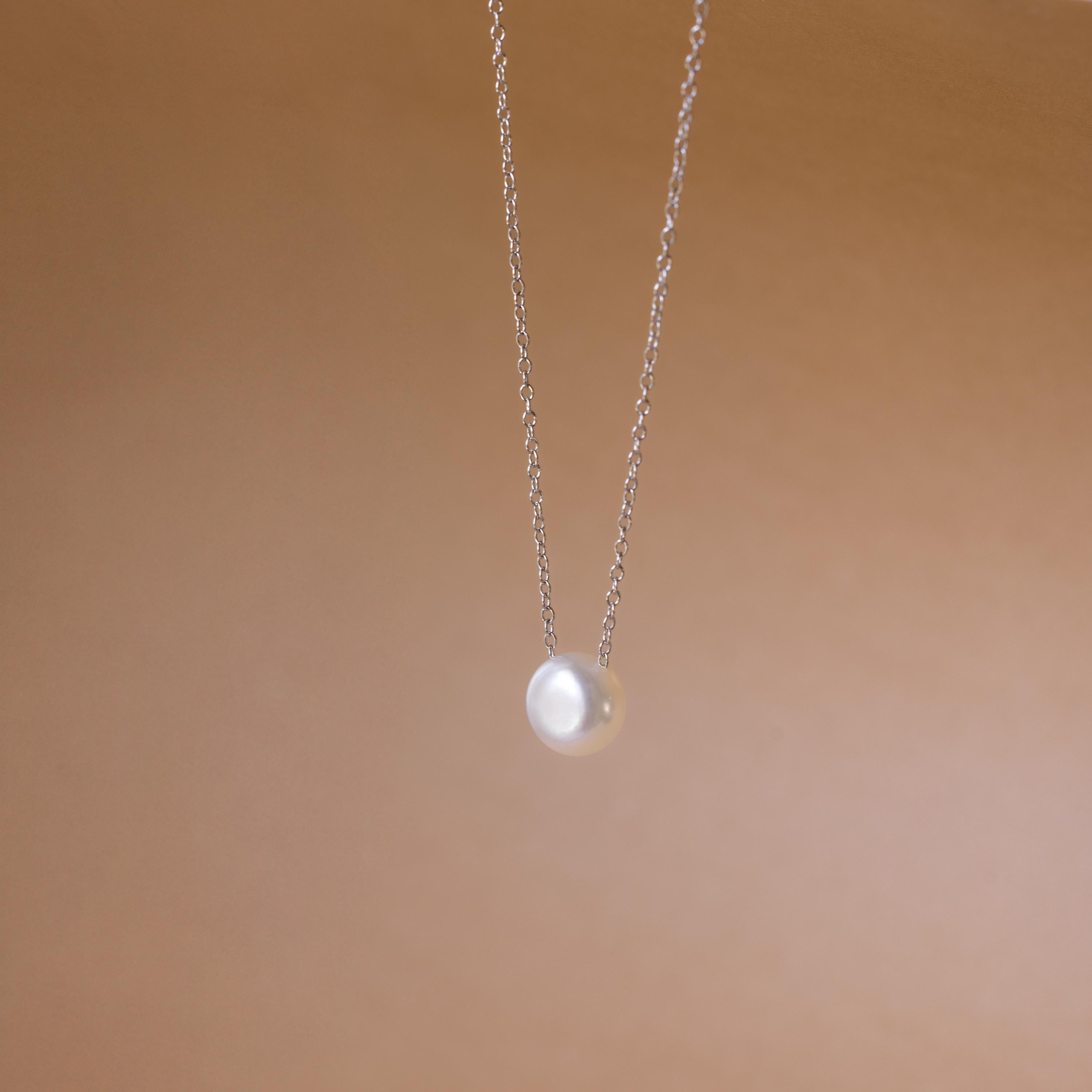 The Classic Pearl Pendant Necklace is a delicate gold chain with a single round pearl, showcased against a soft beige background.