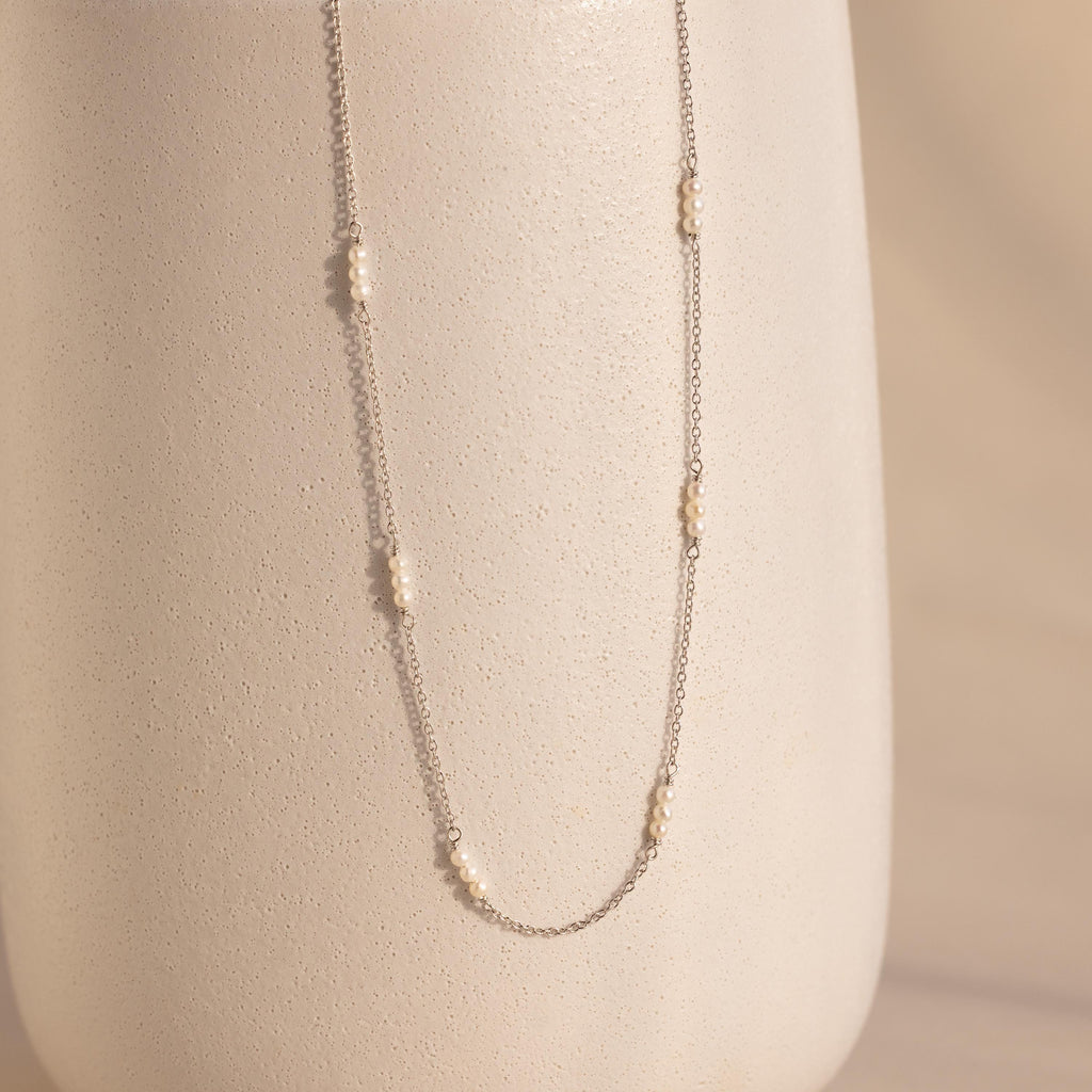 The Hali Pearl Station Necklace features small pearl clusters on a silver chain, shown draped over a textured off-white vase—an elegant, minimalist jewelry piece.