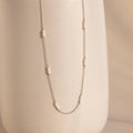The Hali Pearl Station Necklace features small pearl clusters on a silver chain, shown draped over a textured off-white vase—an elegant, minimalist jewelry piece.