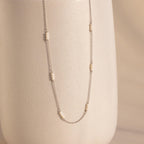 The Hali Pearl Station Necklace features small pearl clusters on a silver chain, shown draped over a textured off-white vase—an elegant, minimalist jewelry piece.