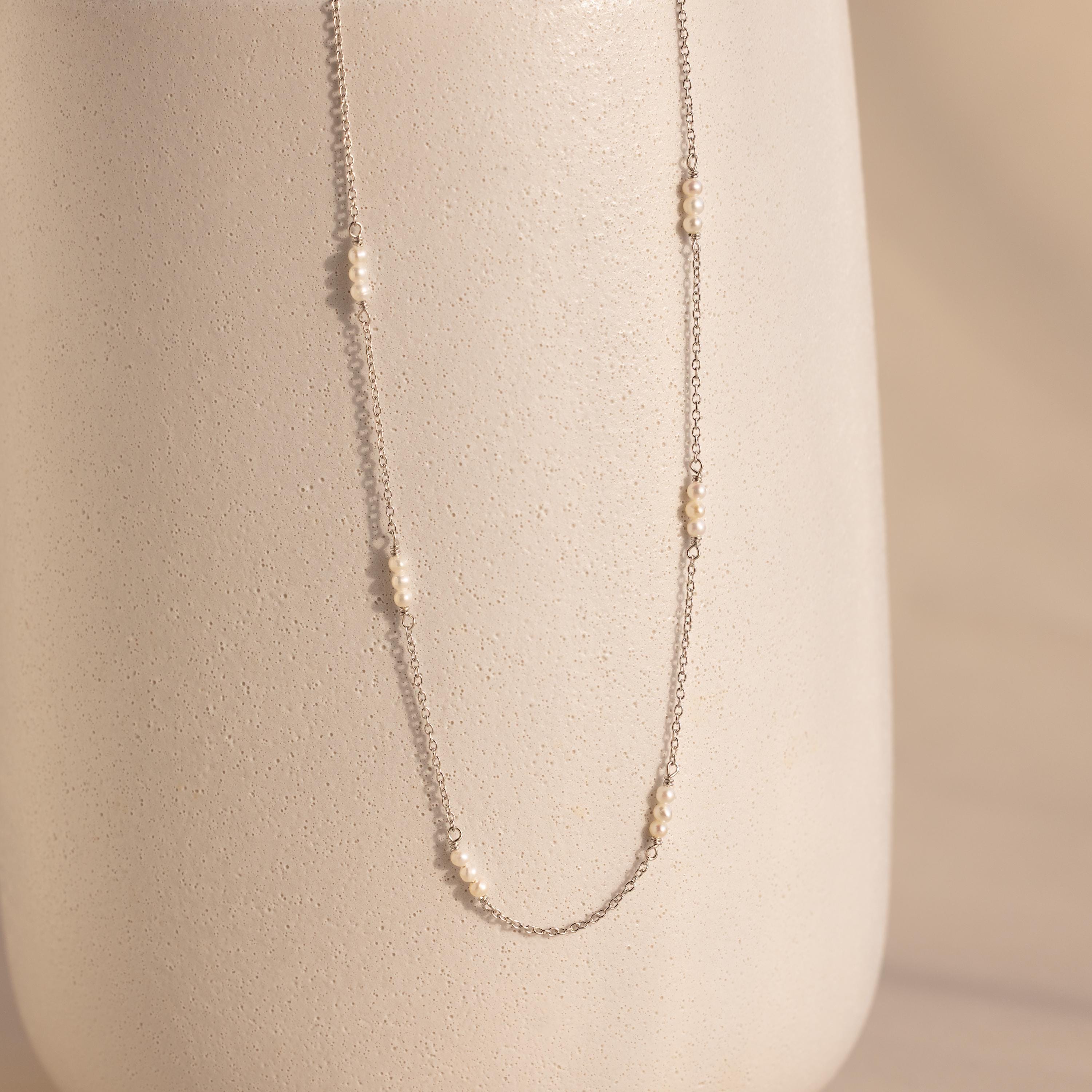 The Hali Pearl Station Necklace features small pearl clusters on a silver chain, shown draped over a textured off-white vase—an elegant, minimalist jewelry piece.