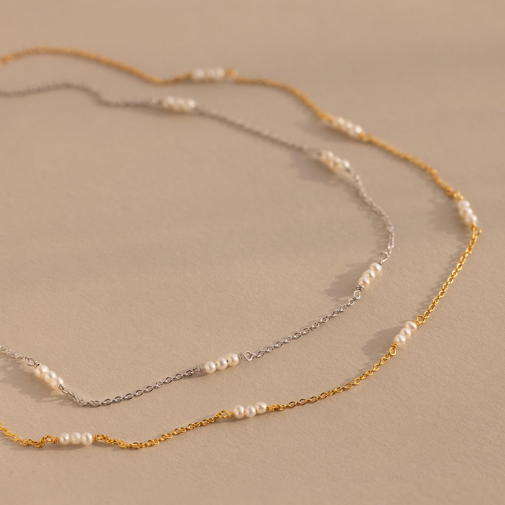 The Hali Pearl Station Necklace, available in silver and gold with small white pearls, highlights minimalist elegance against a beige backdrop.