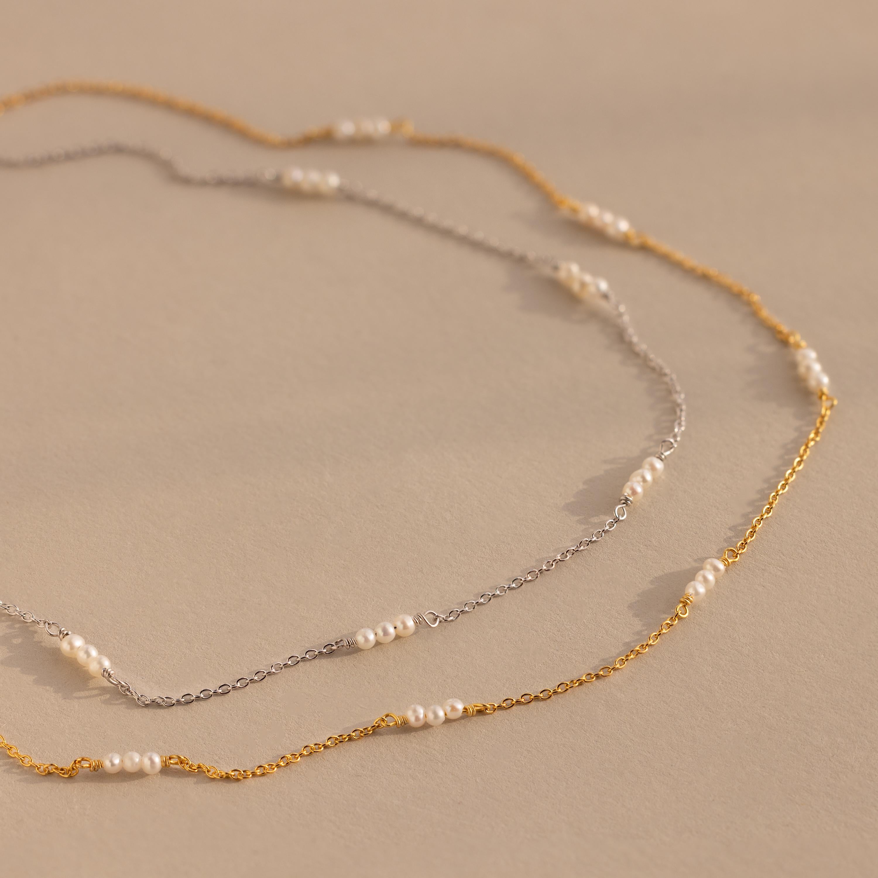 The Hali Pearl Station Necklace, available in silver and gold with small white pearls, highlights minimalist elegance against a beige backdrop.
