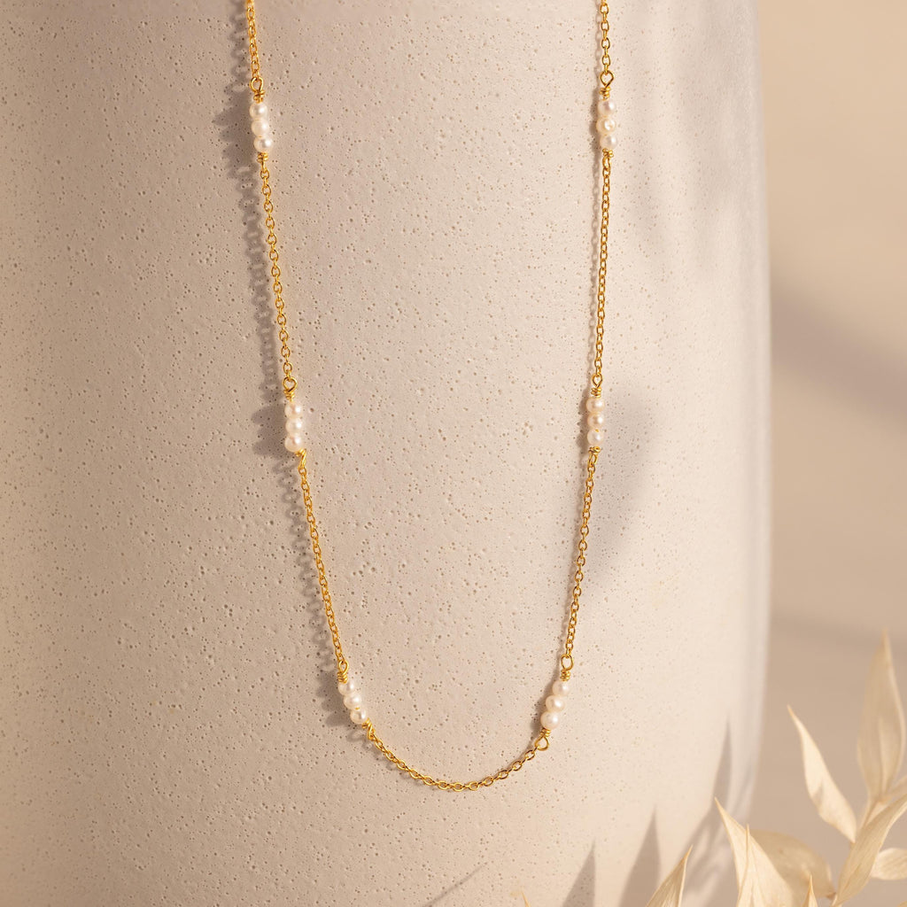 The Hali Pearl Station Necklace features a delicate gold chain adorned with small white pearls, shown on a textured beige background—a minimalist and timeless jewelry piece.