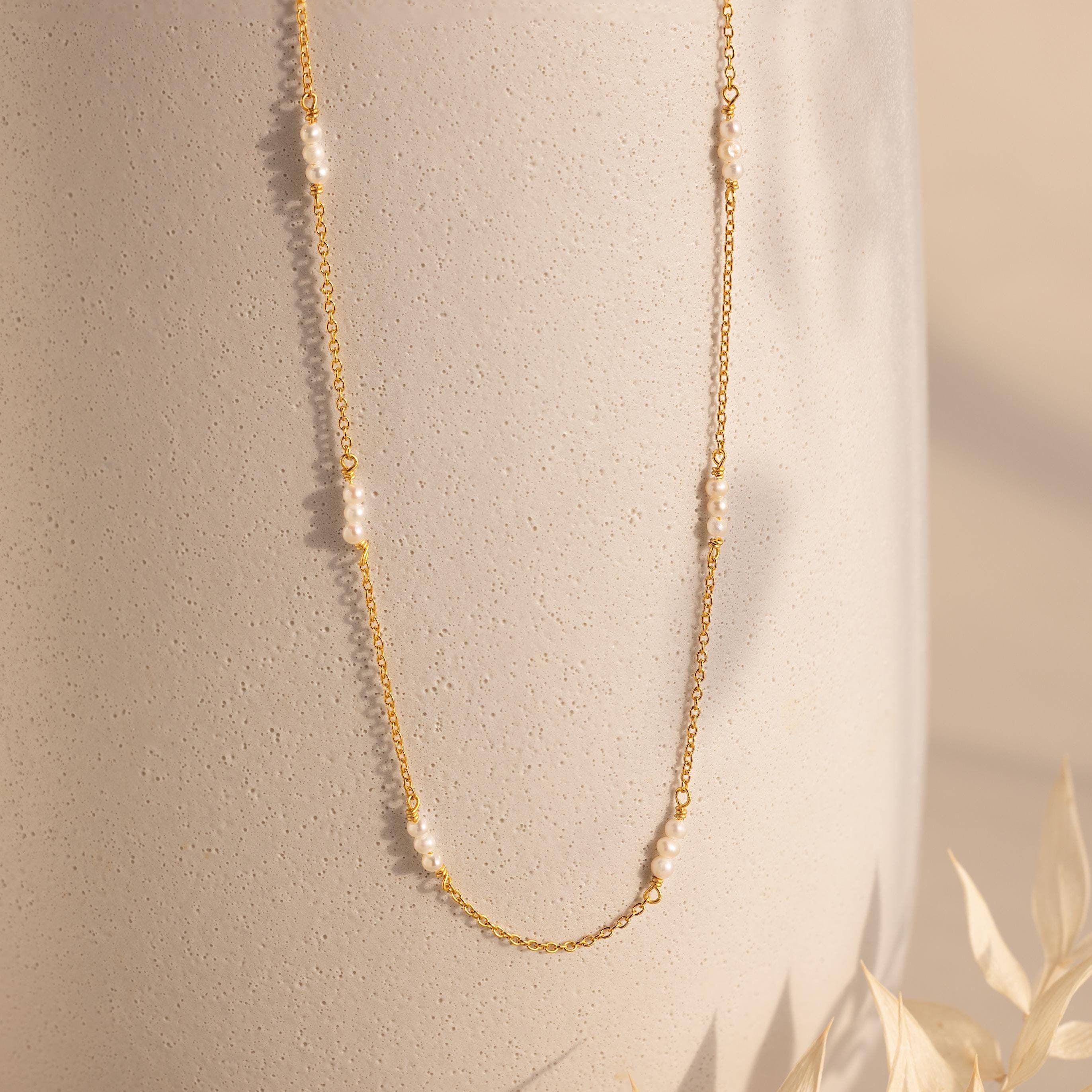 The Hali Pearl Station Necklace features a delicate gold chain adorned with small white pearls, shown on a textured beige background—a minimalist and timeless jewelry piece.