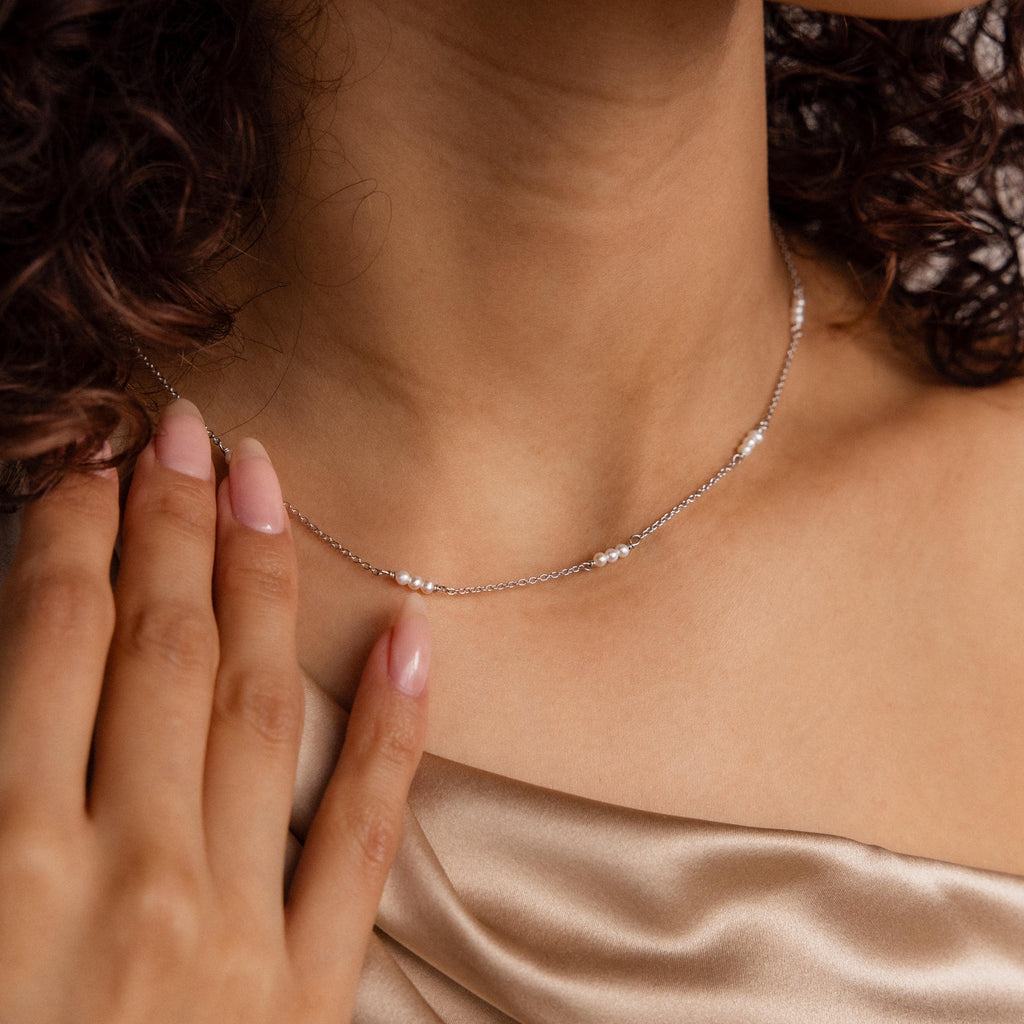 A woman in a champagne satin top gently touching her neck showcases the elegance of minimalist jewelry with the Hali Pearl Station Necklace.