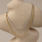 The Oval Beaded Necklace, a gold and silver layered piece, is draped over a textured, abstract beige torso sculpture.