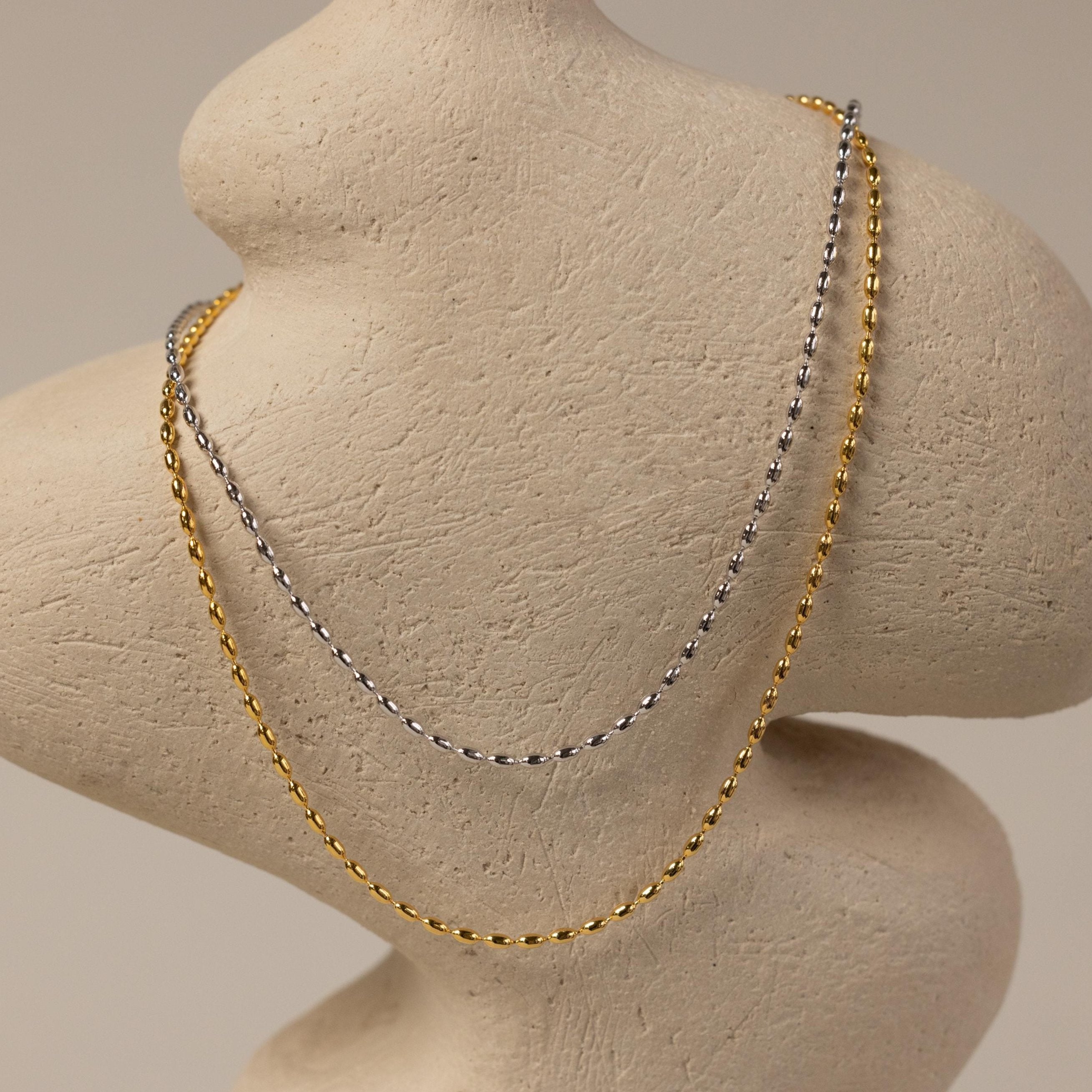 The Oval Beaded Necklace, a gold and silver layered piece, is draped over a textured, abstract beige torso sculpture.