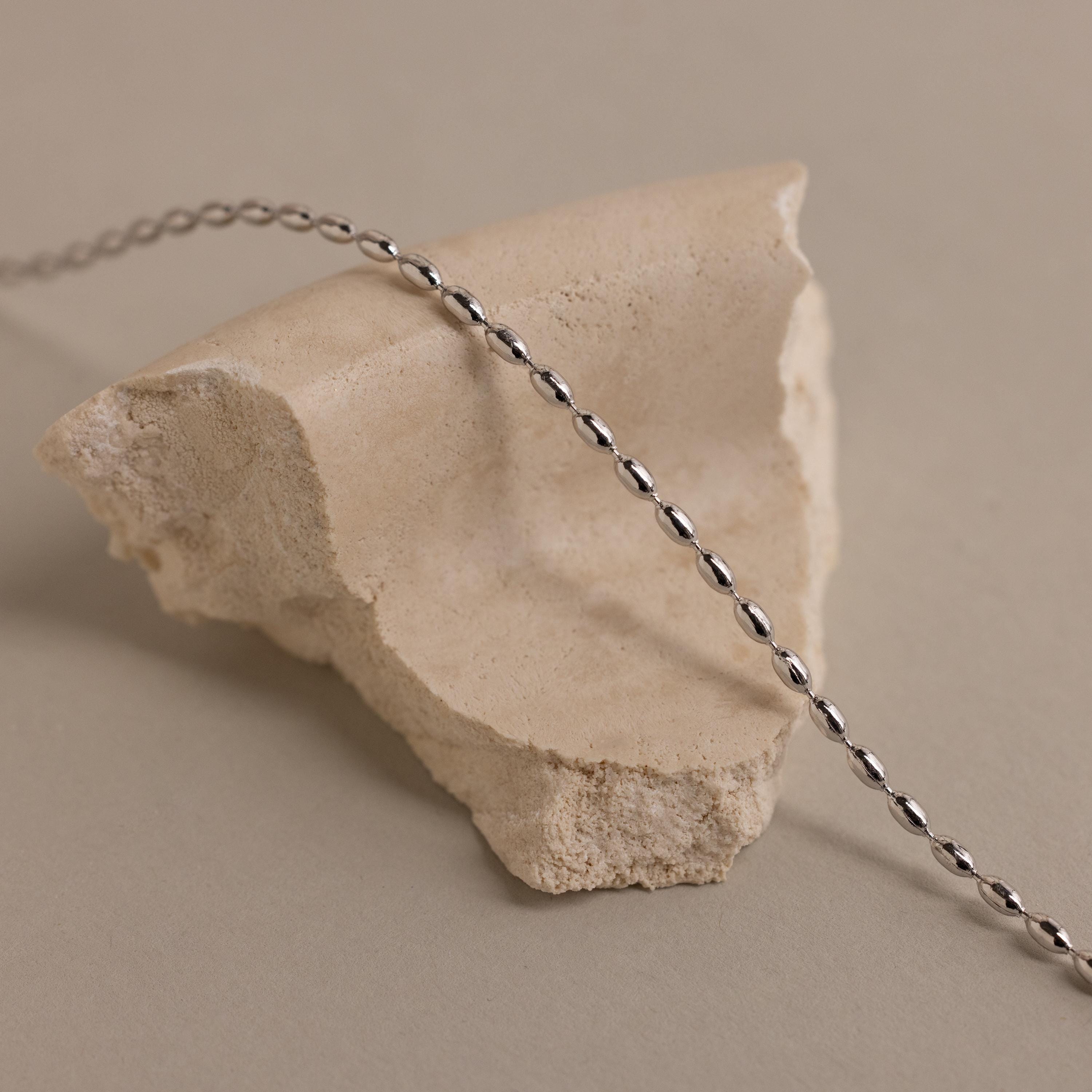 The Oval Beaded Necklace's twisted silver chain is perfect for minimalist wear or layering, shown draped over a rough beige stone on a neutral background.