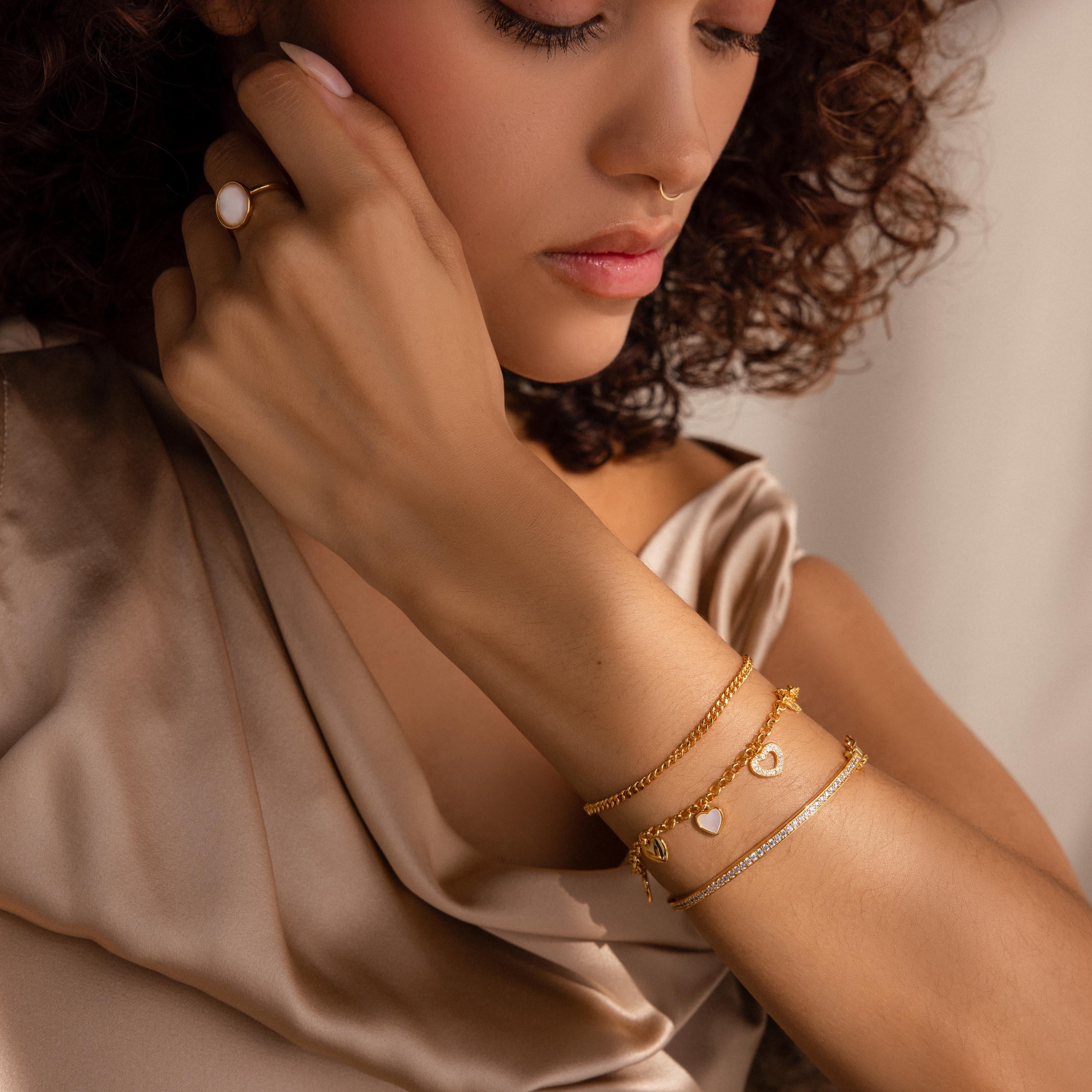 A woman thoughtfully touches her neck, wearing a satin top, a gold ring, and the Heart Charm Bracelet with gold heart charms—a perfect gift for your girlfriend.