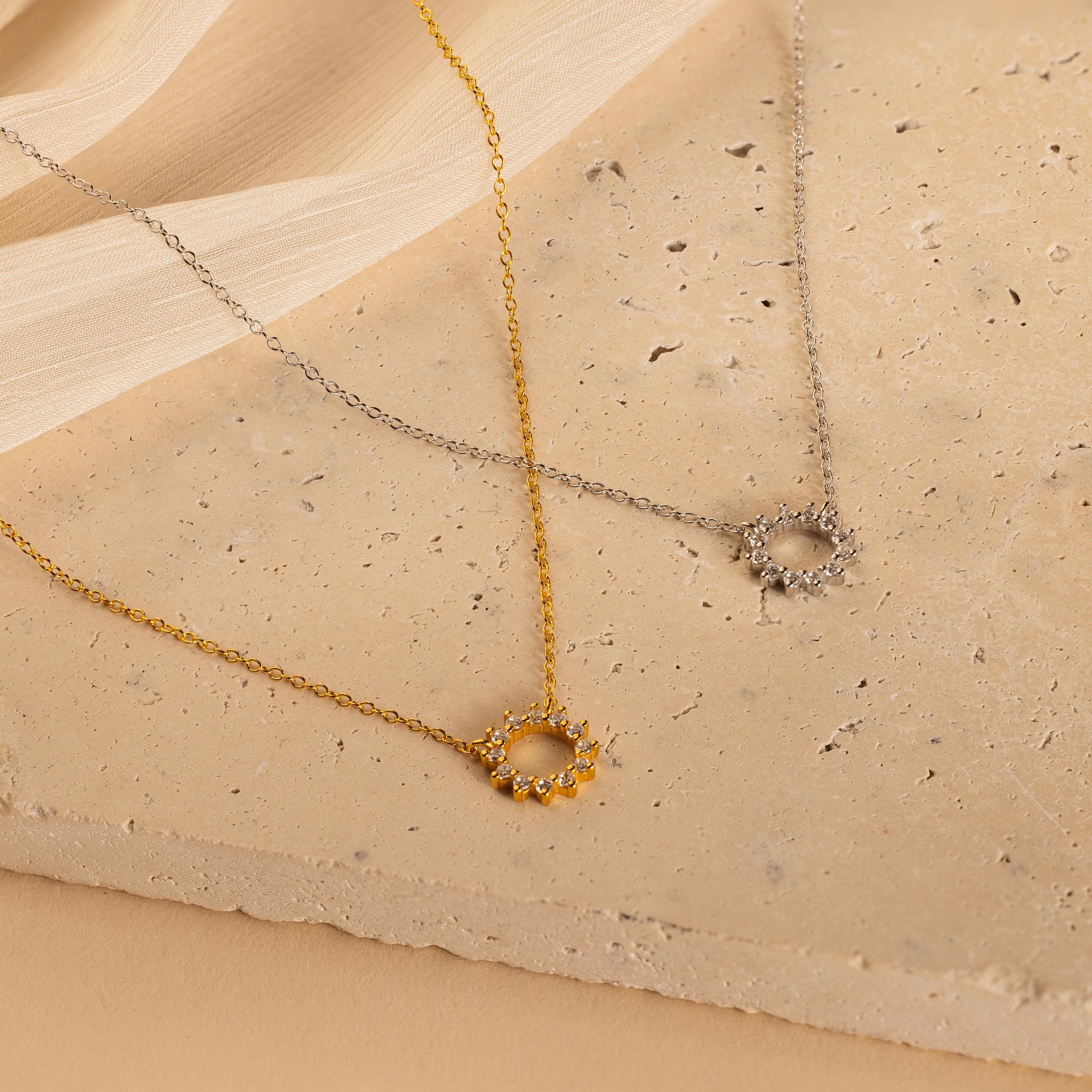 The Diamond Sun Necklace, available in gold and silver, features a delicate sunburst pendant on a textured beige surface, embodying subtle elegance.