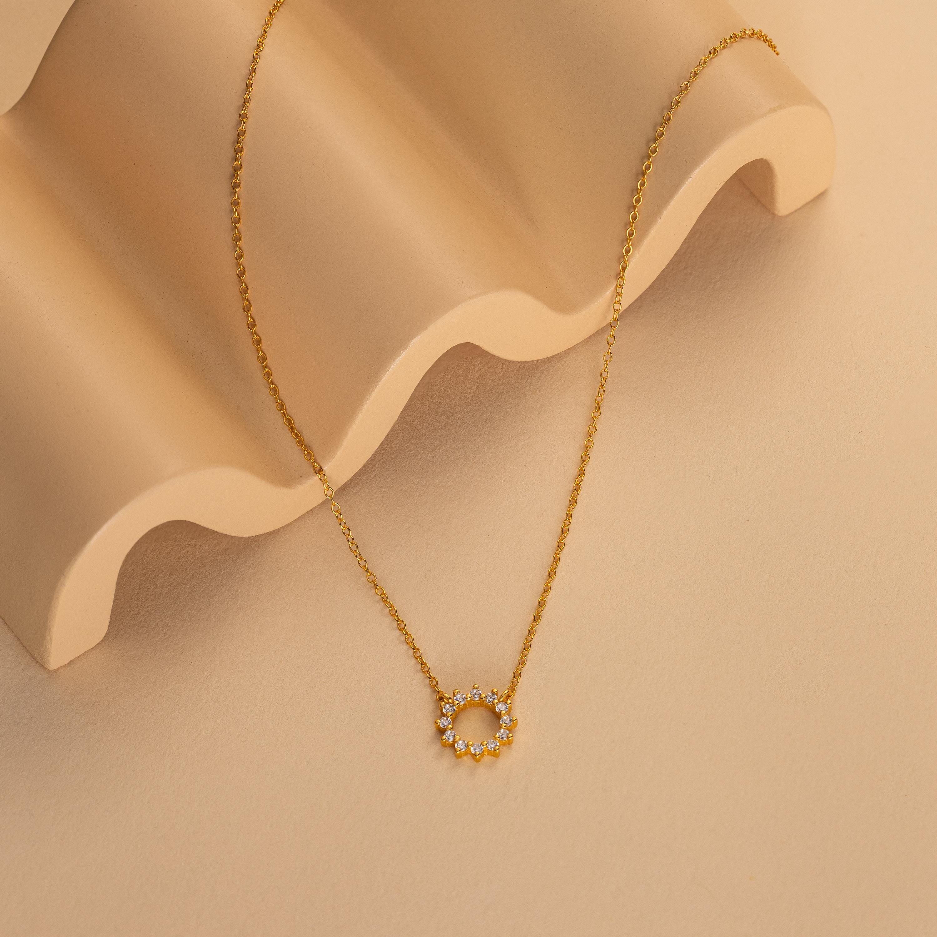 The Diamond Sun Necklace features a circular sunburst pendant adorned with sparkling, diamond-like stones, showcased on a wavy beige surface.