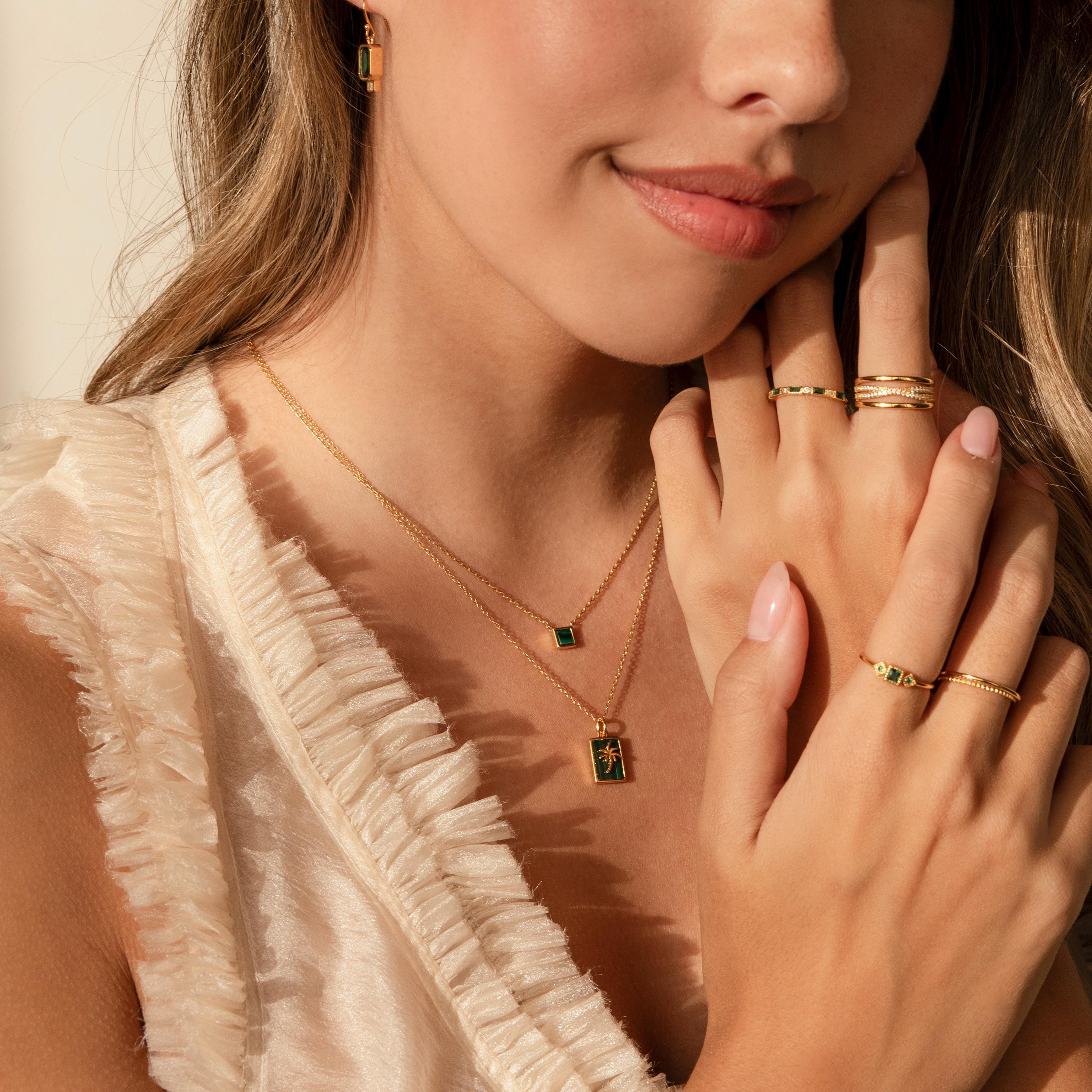 Woman wearing layered gold necklaces, featuring the Malachite Palm Tree Necklace, along with gold rings and earrings, paired with a ruffled cream blouse.