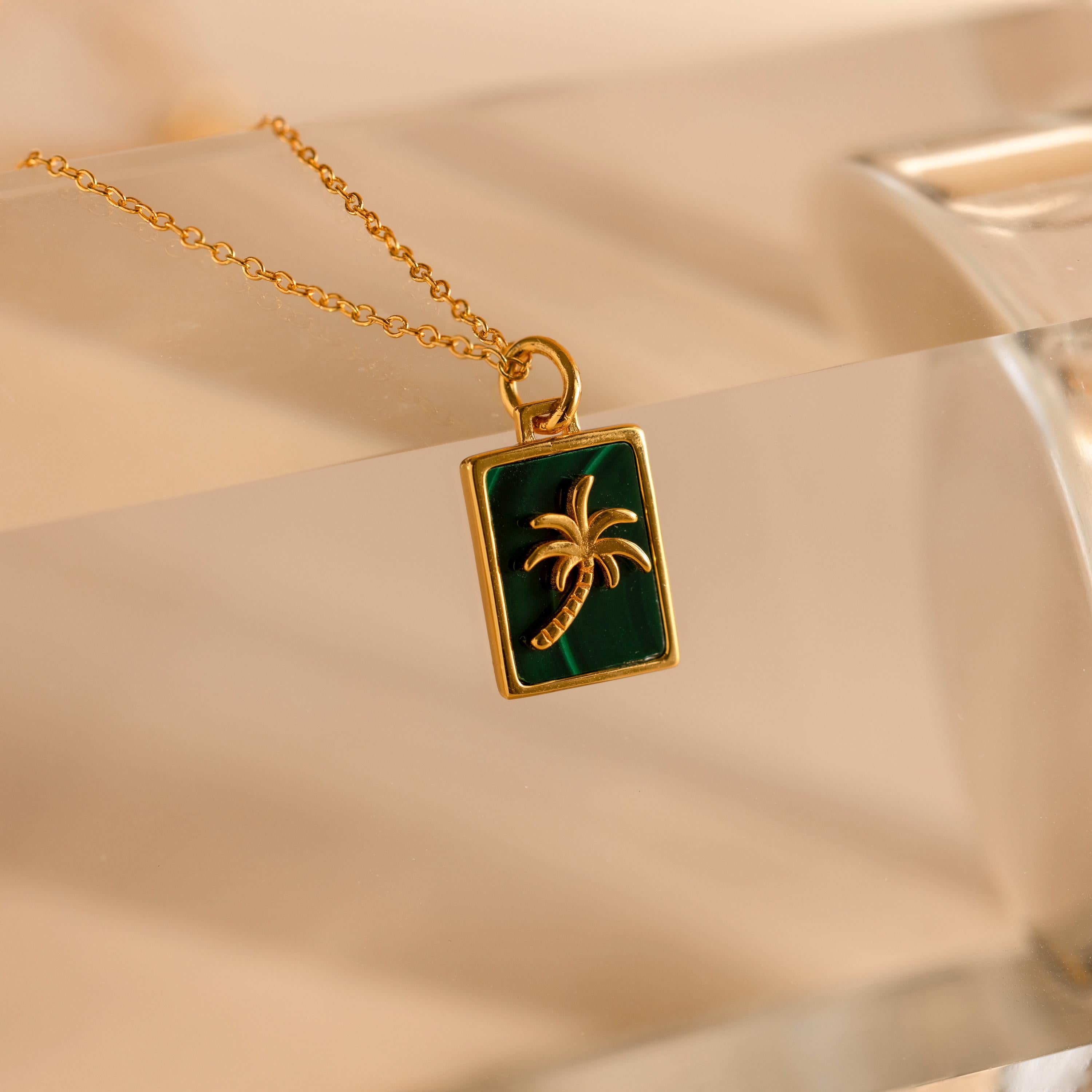 Showcased on a clear surface, the Malachite Palm Tree Necklace features a gold necklace with a rectangular green malachite pendant adorned with a gold palm tree—an elegant choice for palm tree jewelry lovers.