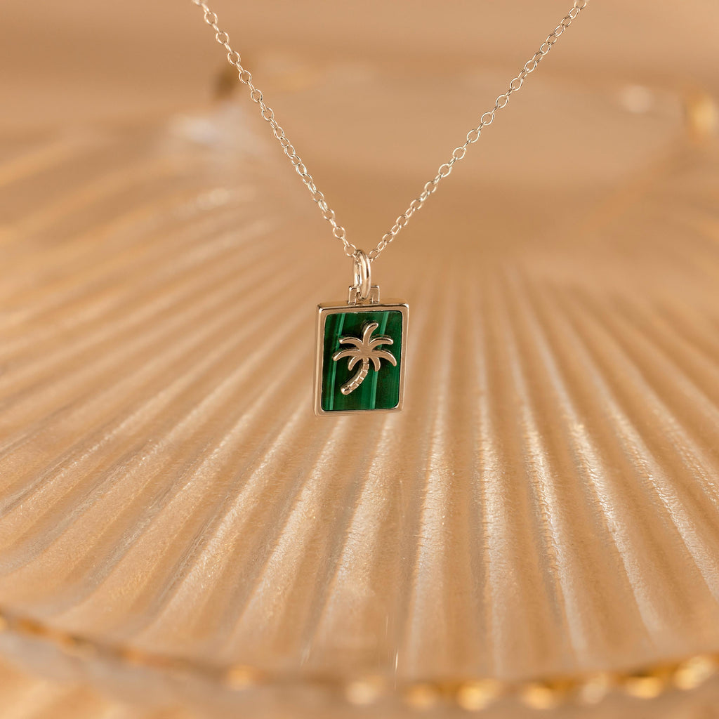 The Malachite Palm Tree Necklace features a green rectangular pendant with a silver palm tree design, displayed on a textured surface.