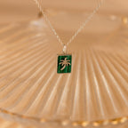 The Malachite Palm Tree Necklace features a green rectangular pendant with a silver palm tree design, displayed on a textured surface.