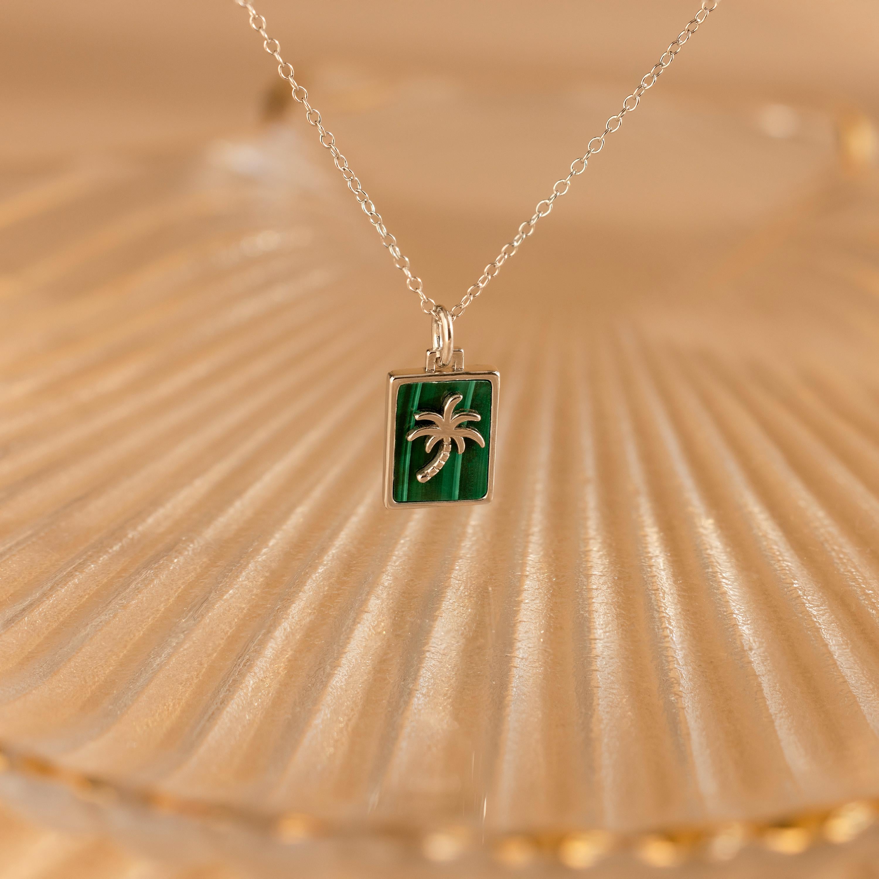 The Malachite Palm Tree Necklace features a green rectangular pendant with a silver palm tree design, displayed on a textured surface.